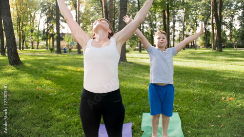 Photo of middle aged female yoga teacher or guru teaching teenage boy doing yoga. Woman with boy meditating and stretching on grass at park
