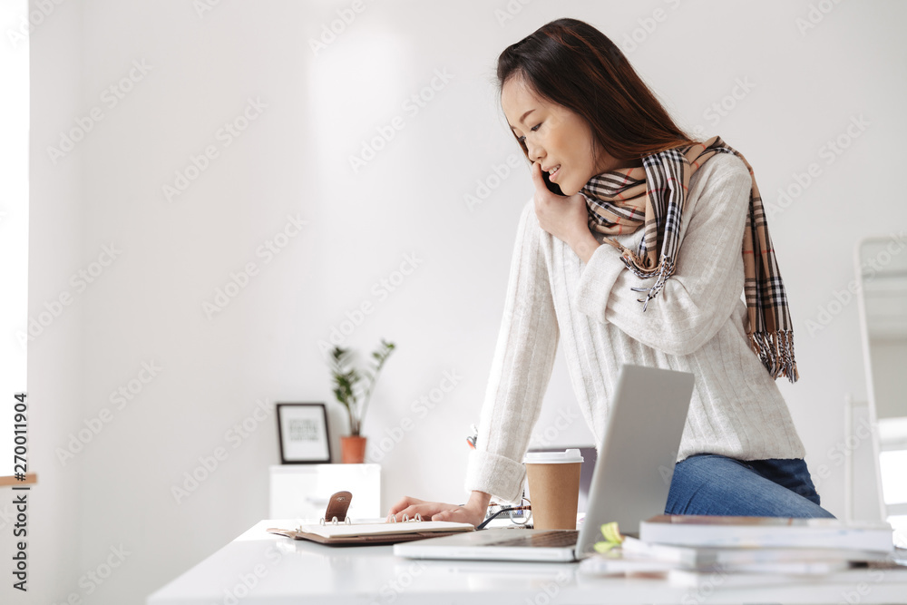 Photo of optimistic asian female worker reading business diary and smiling while sitting on table in office Photo of optimistic asian female worker reading business diary and smiling while sitting on table in office