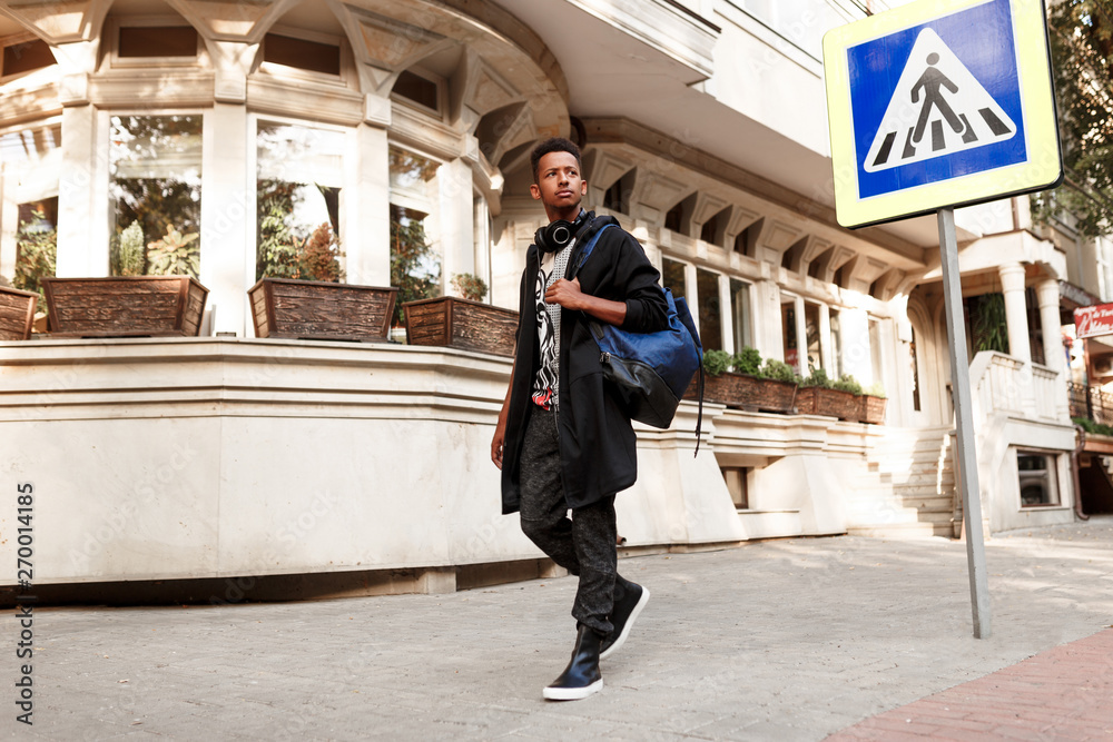 Handsome student outside walking on street, in casual clothes with backpack and headset, near crosswalk, copy space.