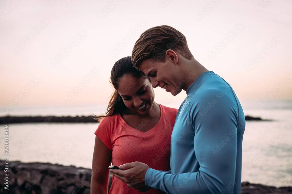 Young couple joggers having a break and using mobile smartphone outdoor - Sporty people having fun with trends technology apps for cellphone - Healthy, tech and youth lifestyle concept