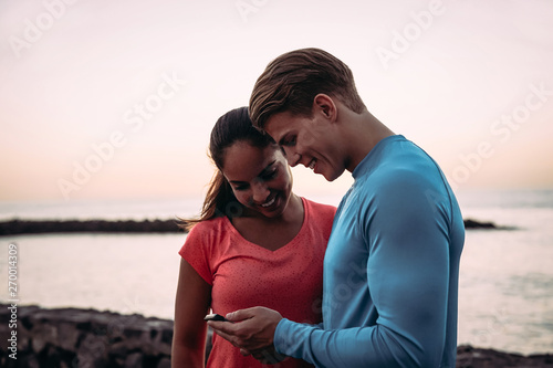Young couple joggers having a break and using mobile smartphone outdoor - Sporty people having fun with trends technology apps for cellphone - Healthy, tech and youth lifestyle concept