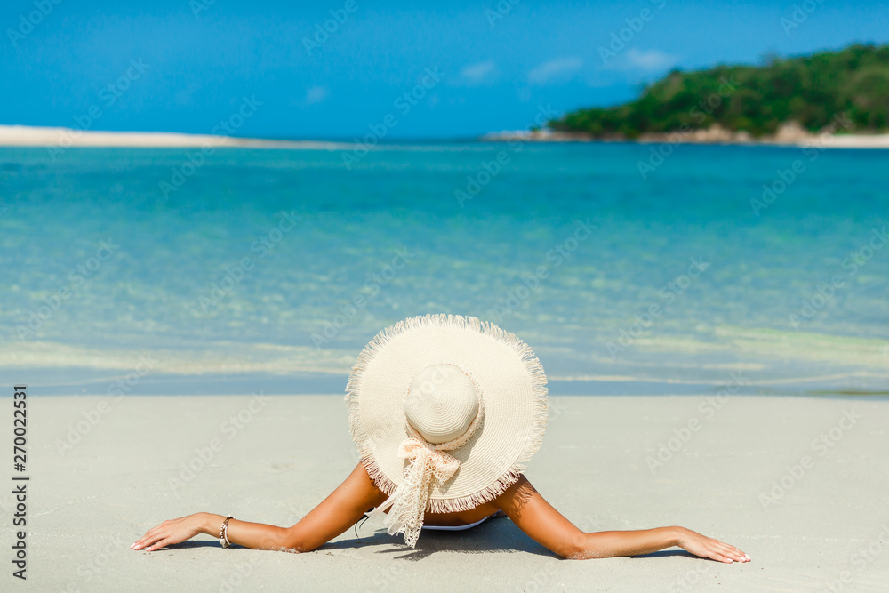 Fototapeta premium Journey. Young beautiful lady in a straw hat, lying on the sand of the seashore with her back to the camera, rest, traveling, concept of perfect state of relaxation and freedom