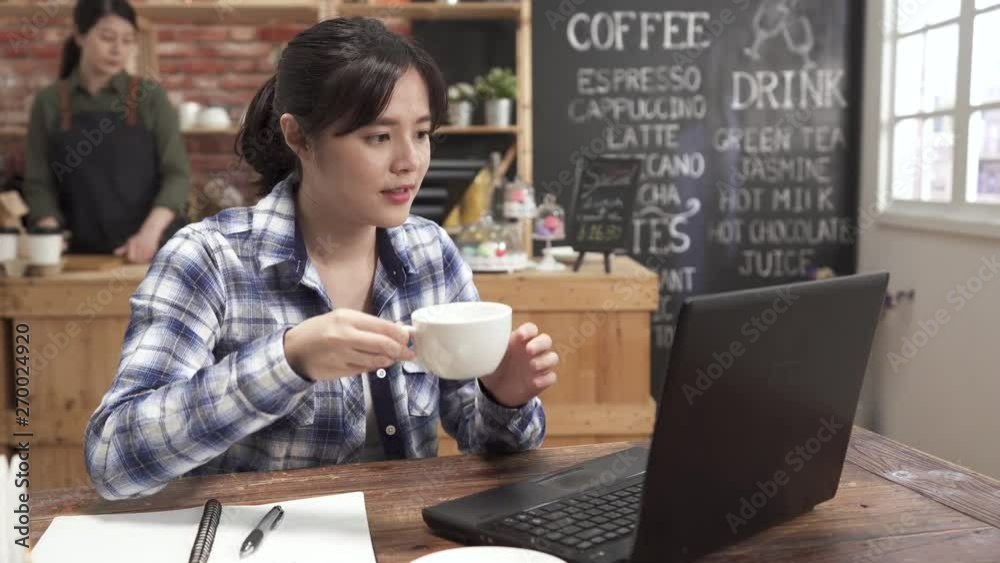 Happy asian japanese woman using laptop chatting online message typing replying smiling at modern coffee shop. busy waitress walking in background back to counter. joy girl drinking cappuccino. Happy asian japanese woman using laptop chatting online message typing replying smiling at modern coffee shop. busy waitress walking in background back to counter. joy girl drinking cappuccino.