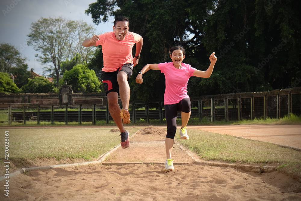 Two sports partners are jogging together on a sunny day wearing orange and pink shirts. They look at each other and smile, enjoy the sports they do