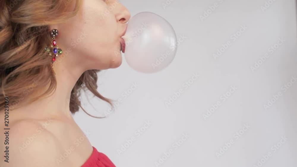 Side view of young woman blowing pink bubble gum on orange isolated background. Closeup female model standing in studio, chewing gum and posing. Concept of burst and refreshment.