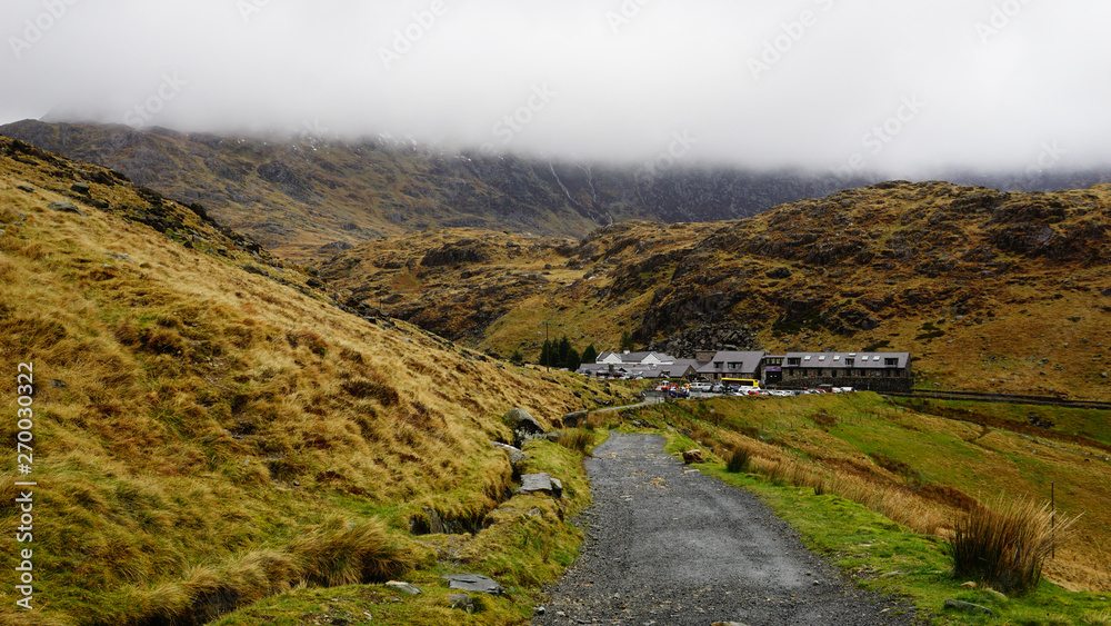 Stone path with saturated grass, beautiful mountains, tiny village houses and a foggy sky – captured during a hike at Snowdon in winter (Snowdonia National Park, Wales, United Kingdom)