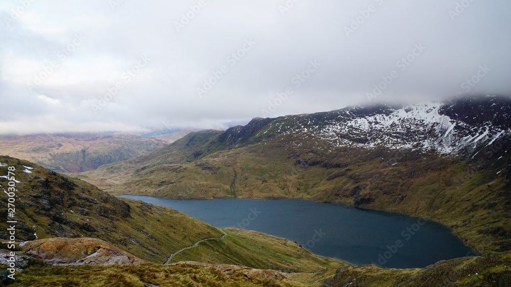 Stunning mountain view with a beautiful lake, foggy sky and some melting snow from the top of the mount – captured during a hike at Snowdon in winter (Snowdonia National Park, Wales, United Kingdom)