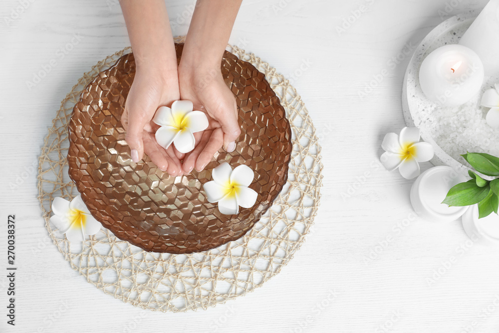 Woman soaking her hands in bowl with water and flowers on wooden table, top view. Spa treatment Woman soaking her hands in bowl with water and flowers on wooden table, top view. Spa treatment