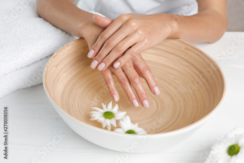 Woman soaking her hands in bowl with water and flowers on table, closeup. Spa treatment