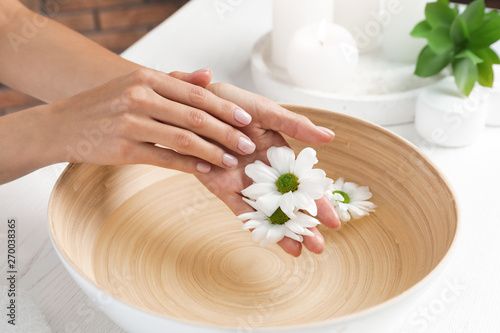 Woman soaking her hands in bowl with water and flowers on table, closeup. Spa treatment