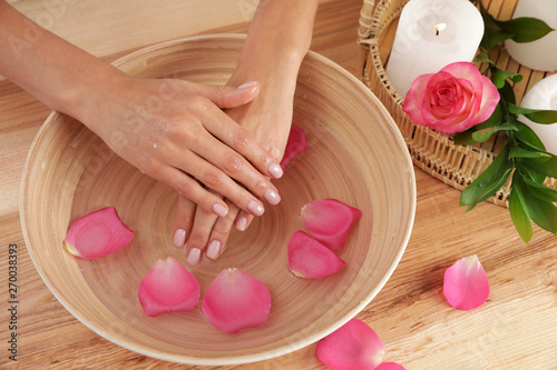 Woman soaking her hands in bowl with water and petals on wooden table, top view. Spa treatment