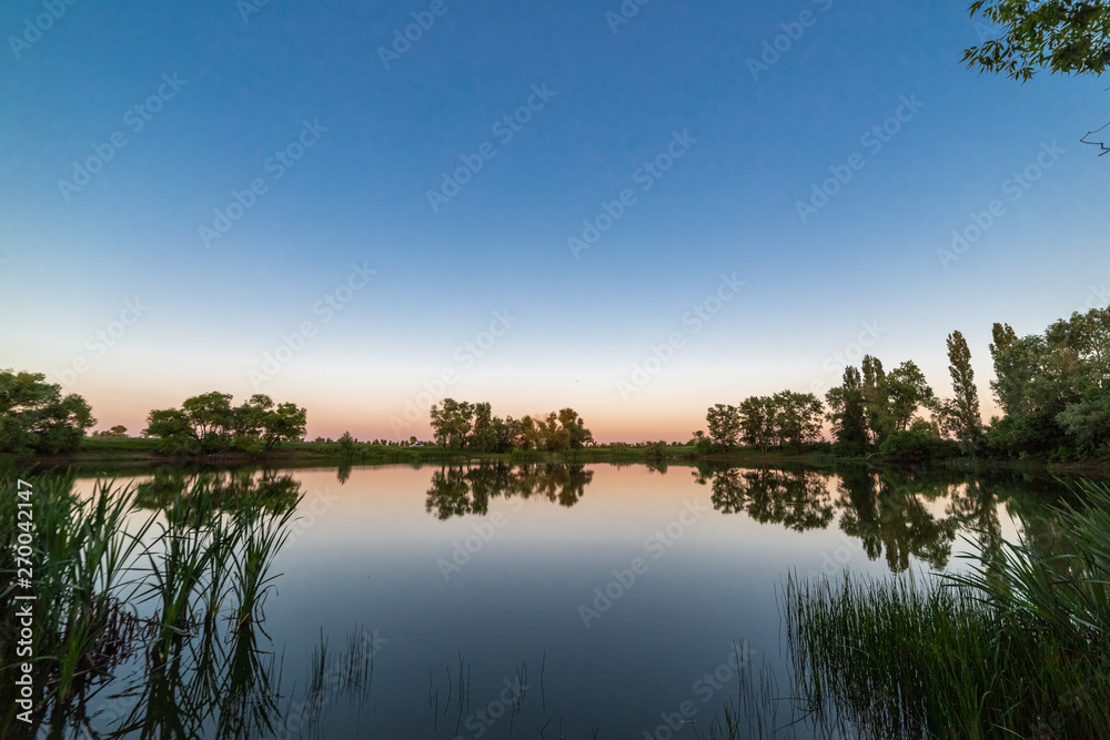 Obraz premium The blue sky and the trees on the shore are reflected in the water at sunset..