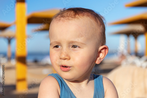 Close up portrait of a cute adorable toddler boy looking aside surprised and charmed.Llittle funny baby child is sitting on the beach of the tropical resort in summer noon.