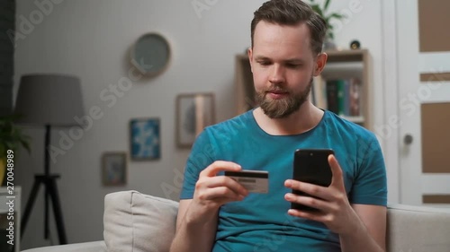 Young caucasian man sitting on the couch, using smart phone for online shopping with credit card on living room. Indoor. Guy holding credit card, easy pay by using digital mobile device app.