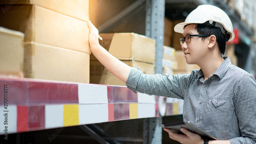 Young Asian man worker wearing safety helmet and eyeglasses doing stocktaking of product in cardboard box on shelves in warehouse by using digital tablet and pen. Physical inventory count concept