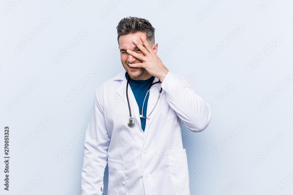 Young handsome doctor man blink at the camera through fingers, embarrassed covering face. Young handsome doctor man blink at the camera through fingers, embarrassed covering face.