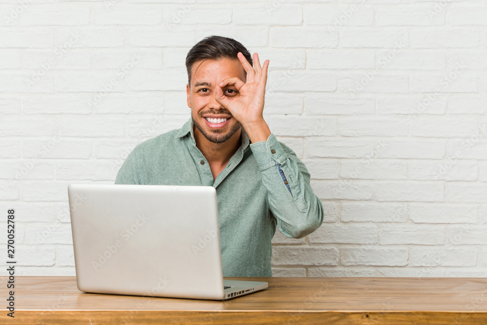 Young filipino man sitting working with his laptop excited keeping ok gesture on eye. Young filipino man sitting working with his laptop excited keeping ok gesture on eye.