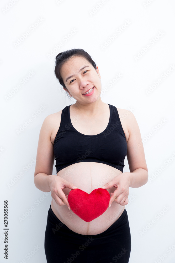Young beautiful Asian pregnant woman touching her belly at home and smiling dressed in spandex dress with hands holding red heart isolated on white background Young beautiful Asian pregnant woman touching her belly at home and smiling dressed in spandex dress with hands holding red heart isolated on white background