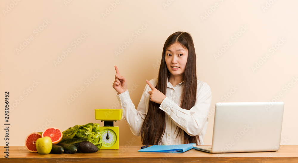 Young nutritionist chinese woman working with her laptop shocked pointing with index fingers to a copy space. Young nutritionist chinese woman working with her laptop shocked pointing with index fingers to a copy space.