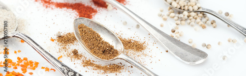 panoramic shot of various scattered spices in silver spoons on white background