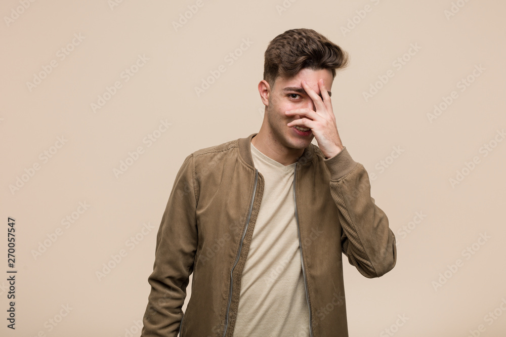 Young caucasian man wearing a brown jacket blink at the camera through fingers, embarrassed covering face. Young caucasian man wearing a brown jacket blink at the camera through fingers, embarrassed covering face.