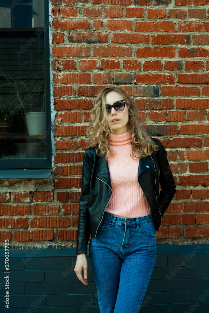 blonde girl with curly hair in black glasses, pink turtleneck, in a black leather rocker jacket on the background of a red brick wall, with a place for the inscription on the left