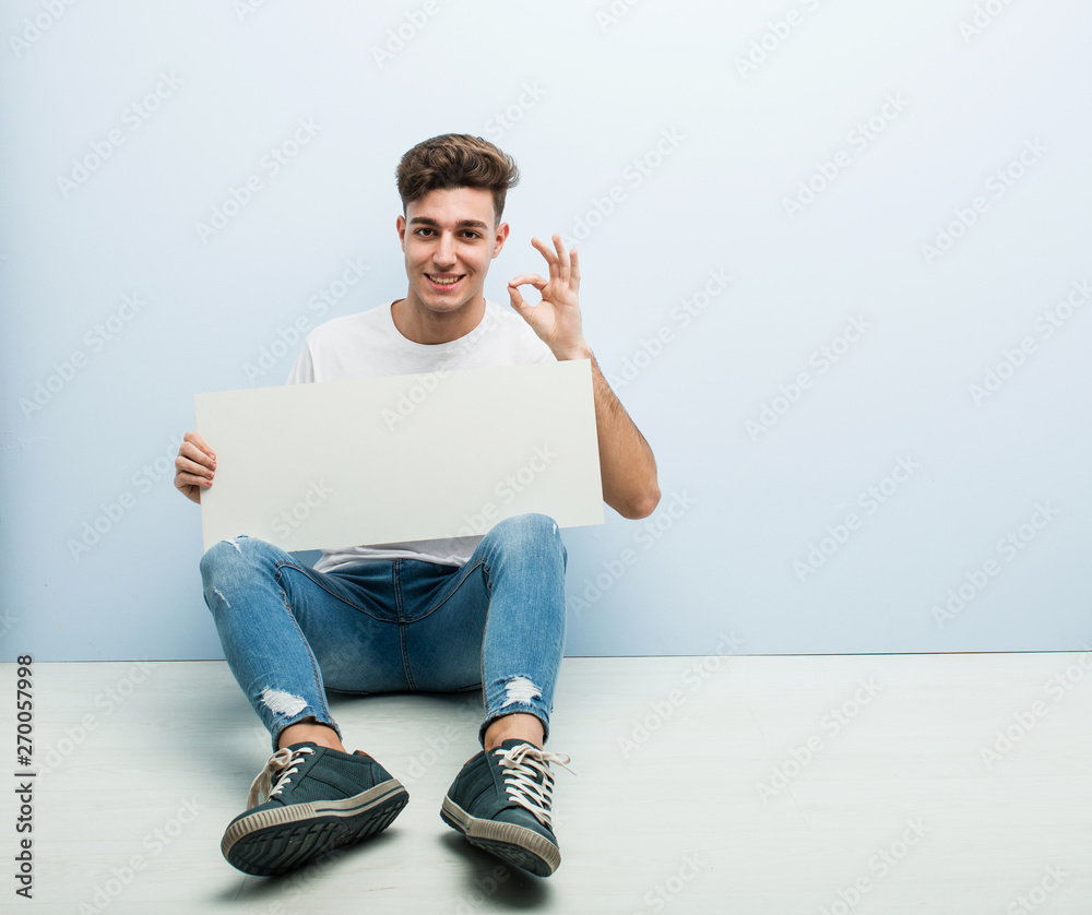 Young man holding a placard sitting on his home floor cheerful and confident showing ok gesture. Young man holding a placard sitting on his home floor cheerful and confident showing ok gesture.