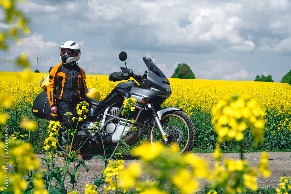 Girl in a protection outfit and glasses with touristic motorcycle. Rape yellow flowers field on background. Adventure trail tour, enduro and off road, summer day. extreme vacation concept