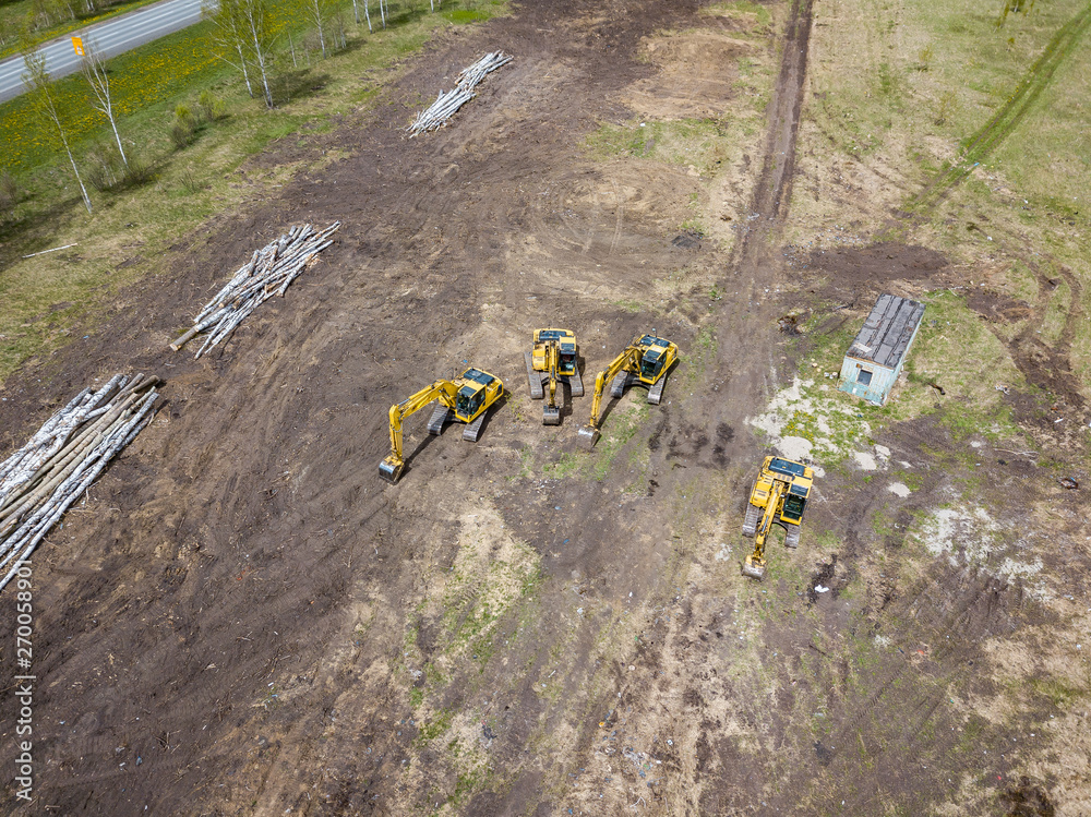Naklejka premium Aerial top view of four yellow crawler excavators standing on ground near the construction site and waiting for the working day to start digging the ground near the building, road in field among tree