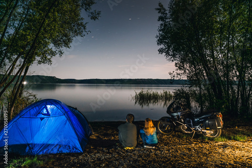 Night camping on lake shore. Man and woman is sitting. Couple tourists enjoying amazing view of night sky full of stars. Blue tent  adventure motorcycle  active lifestyle. Bikers  motorcyclist