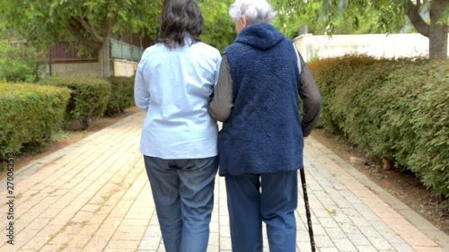 Old woman with walking stick strolling with companion or female caregiver in a green alley. They go away from a camera / from focus to blur/shooting from the back. Shallow depth of field.