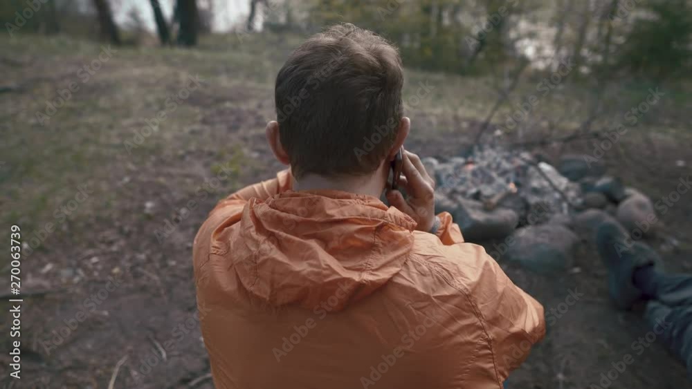Tourist hiker talking on the phone at the campfire enjoying the warmth of the fire in nature on a summer evening. Camping tourists during a hike in the woods. Tourist hiker talking on the phone at the campfire enjoying the warmth of the fire in nature on a summer evening. Camping tourists during a hike in the woods.