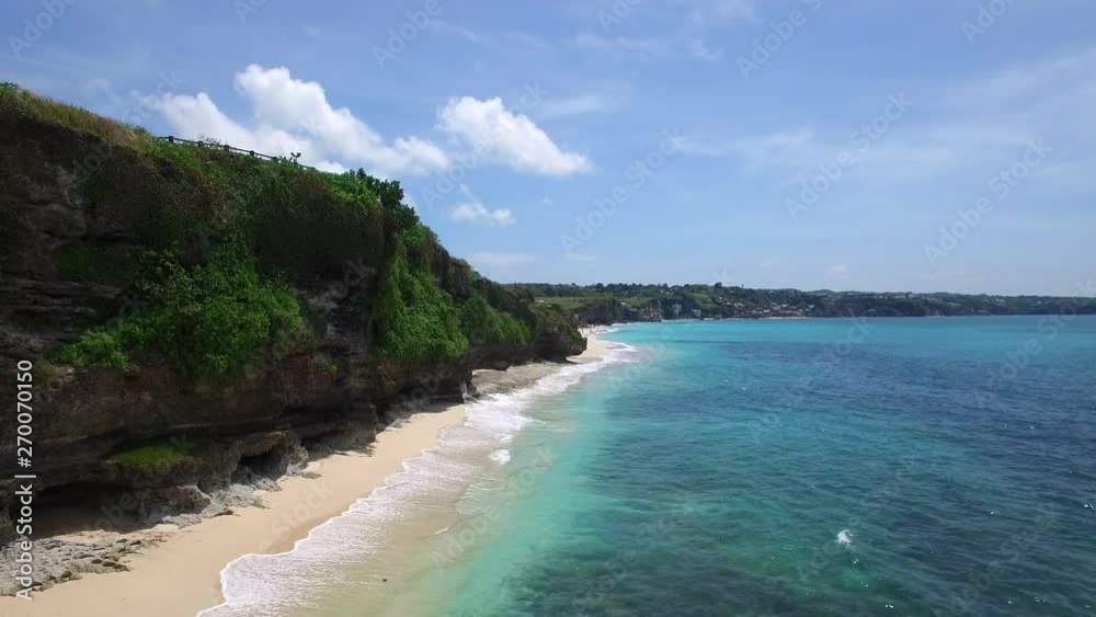 Long sandy beach on tropical island surrounded by cliffs with green vegetation on top and azure Indian Ocean with frothy waves on bright sunny day. Aerial footage of Dreamland beach, Bali, Indonesia. Long sandy beach on tropical island surrounded by cliffs with green vegetation on top and azure Indian Ocean with frothy waves on bright sunny day. Aerial footage of Dreamland beach, Bali, Indonesia.