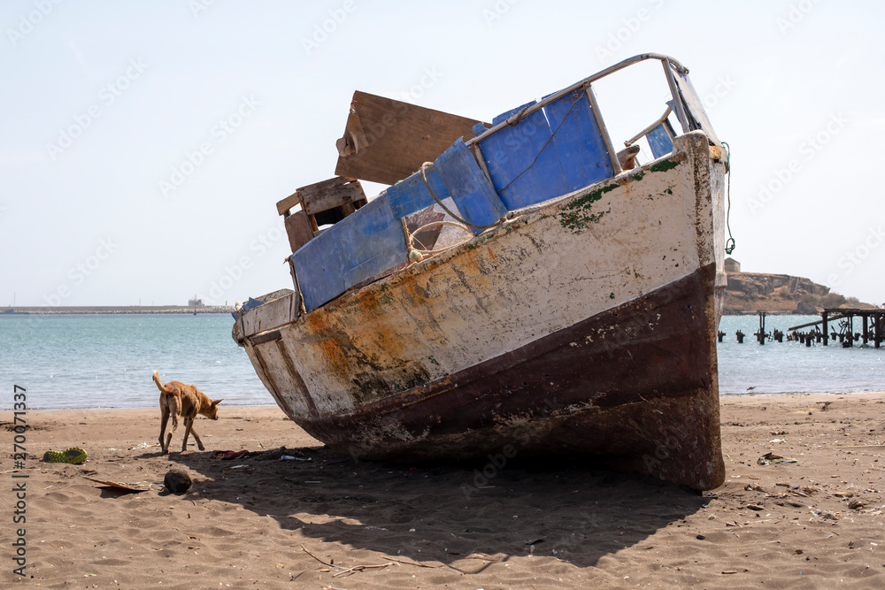Stray dog ​​walks next to an old crooked fishing boat, on the city beach of Praia, island of Santiago, Cape Verde, Cabo Verde.