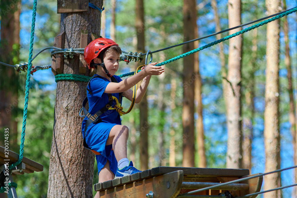 Young child boy in safety harness and helmet attached with carbine to cable moves confidently along rope way in recreation park.  Sport, game, leisure concept.