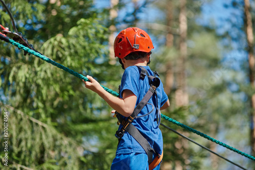 Child in forest adventure park. Kid in red helmet and blue t- shirt climbs on high rope trail. Agility skills and climbing outdoor amusement center for children.