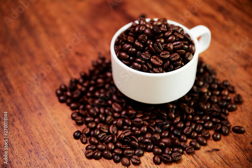 Close-up of white cup with heap of aroma coffee beans in it and near on wooden background