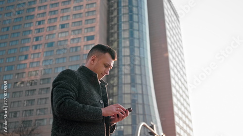 Adult Caucasian Confident Young Business Man is Using Smartphone App and Walking Outside near Modern Office Building in the City Center. Medium Low Angle