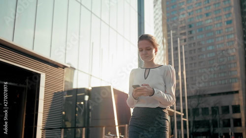 Adult Caucasian Confident Young Business Woman with Hair Bun is Using Smartphone App and Walking Outside near Modern Office Building. Medium Long Low Angle