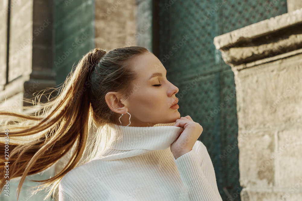 Fashionable woman with long hair in ponytail hairstyle wearing trendy hoop  earrings, white turtleneck sweater, posing in street of European city.  Copy, empty space for text Stock Photo | Adobe Stock