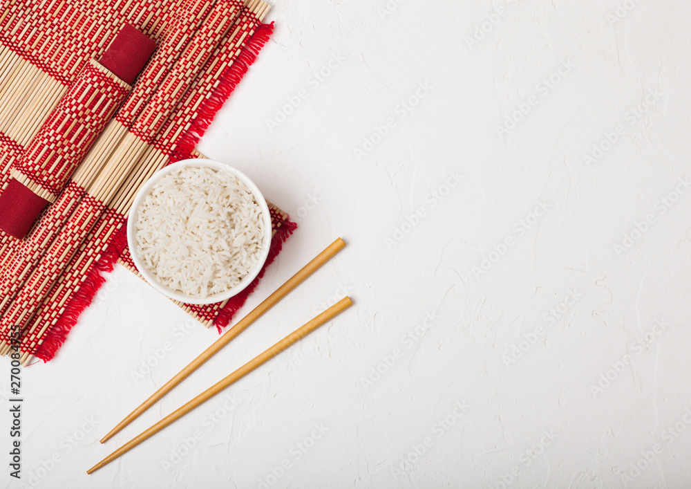 White bowl with boiled organic basmati jasmine rice with wooden chopsticks and sweet soy sauce on bamboo placemat with red linen towel on black stone background. White bowl with boiled organic basmati jasmine rice with wooden chopsticks and sweet soy sauce on bamboo placemat with red linen towel on black stone background.