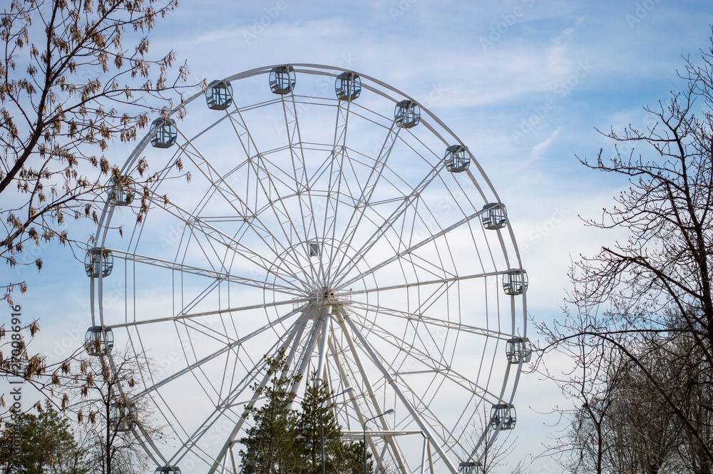 White Ferris wheel with glass light-blue cabins against the blue sky in trees frame. Wheel in amusement city park. White Ferris wheel with glass light-blue cabins against the blue sky in trees frame. Wheel in amusement city park.
