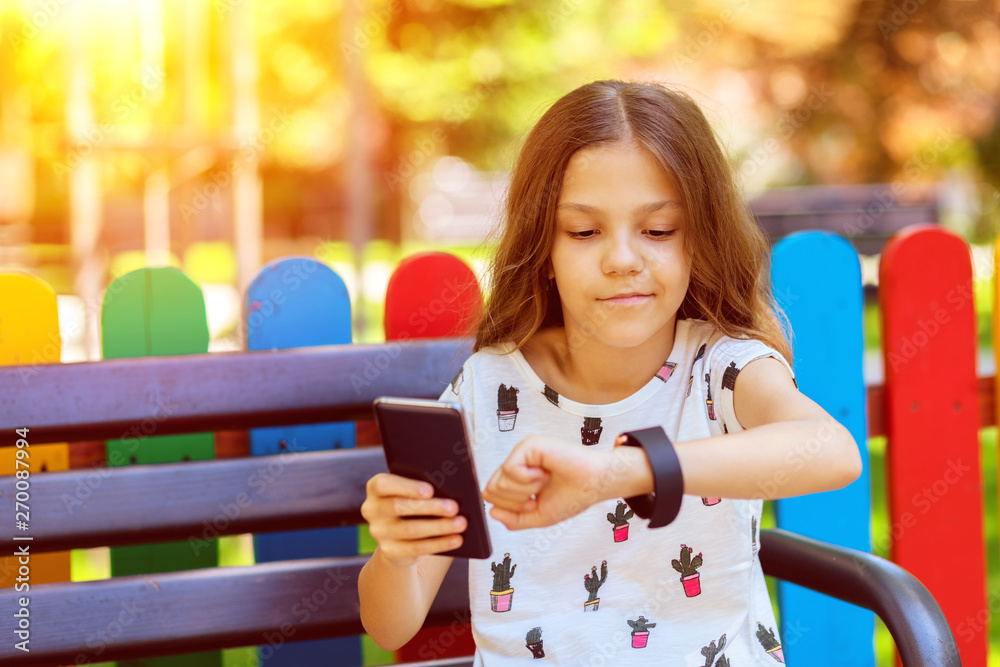 Smiling little girl using modern wearable smart watch and mobile phone  outdoor synching data – happy kid with trendy gadgets synchronizing  smartwatch with smartphone while sitting on bench in park Stock Photo