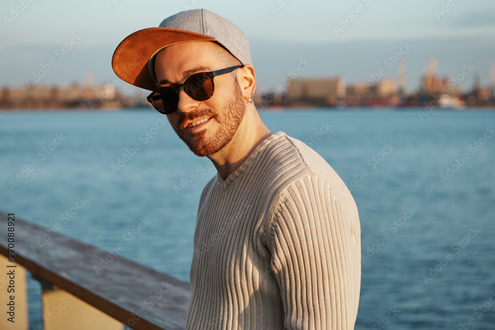 Urban lifestyle, leisure, city, people and fashion concept. Picture of cute handsome young man with neat trimmed beard and mustache posing at wide river with blurred buildings in background