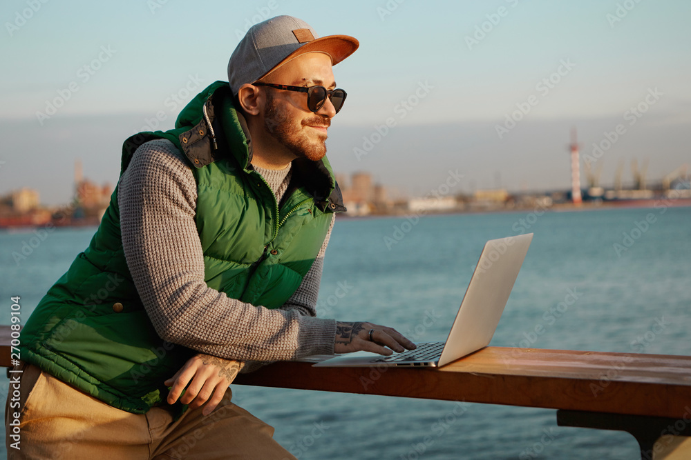 People, inspiration, modern lifestyle and electronic devices concept. Side view of stylish young unshaven man blogger in snapback and black shades using laptop for remote work, creating new content