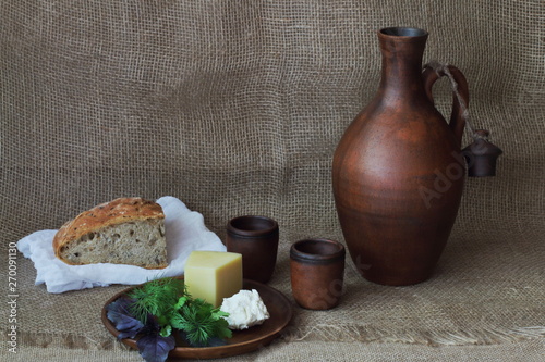 Hard and soft cheese with greens on round clay plate, jug for wine, cup and bread on white cloth on homespun rough background. Concept — natural food, rustic style, pottery.