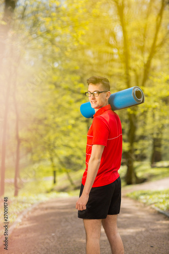 Young sports man in red t-shirt standing on the park track and posing with a blue yoga mat. Behind it a blurred background