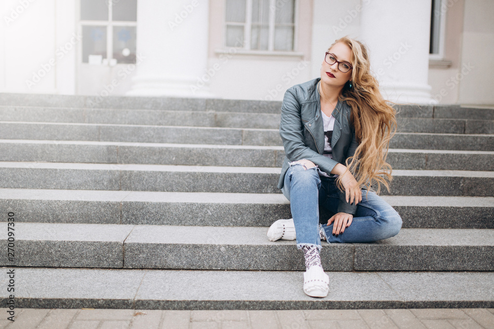 Close up street urban portrait of young fashion stylish pretty blondie woman in fashion jeans in glasses sitting outdoors in the street . Beauty, student, freedom concept