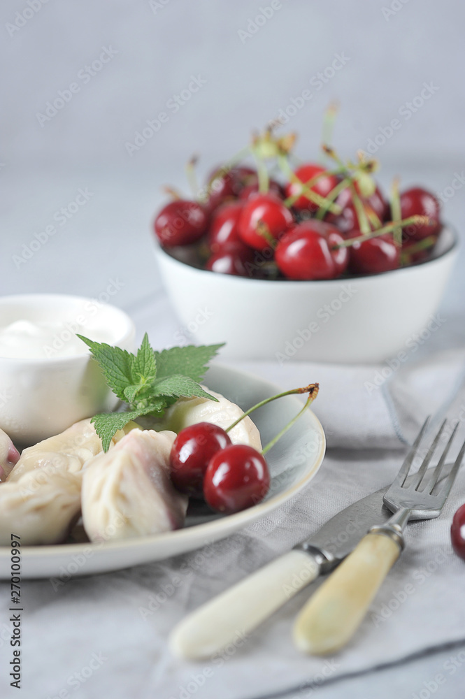 Dumplings with cherries and sour cream. A cup of cherries, cutlery and a napkin complement the composition. Light background. Vertical frame orientation. Dumplings with cherries and sour cream. A cup of cherries, cutlery and a napkin complement the composition. Light background. Vertical frame orientation.
