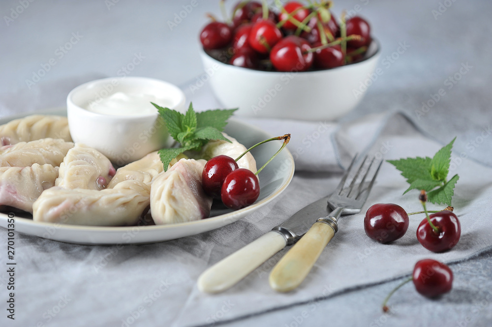 Dumplings with cherries and sour cream. A cup of cherries, cutlery and a napkin complement the composition. Light background. Dumplings with cherries and sour cream. A cup of cherries, cutlery and a napkin complement the composition. Light background.